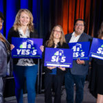 Panel of Conference speakers holding prop 5 signs