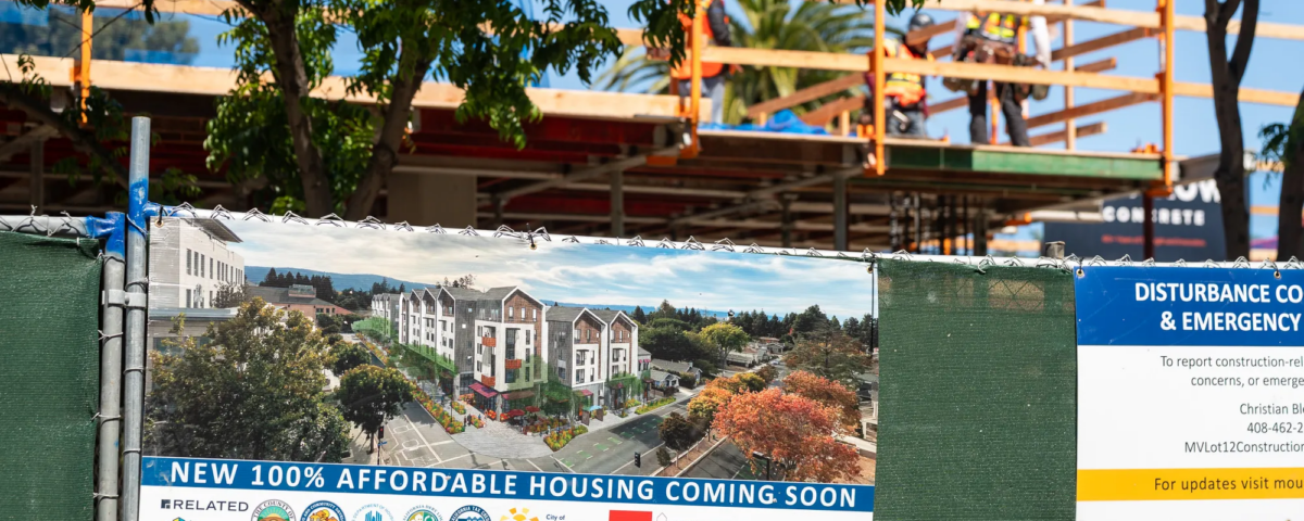 Green fence with sign that signifies affordable housing is being built. Workers in the background out of focus
