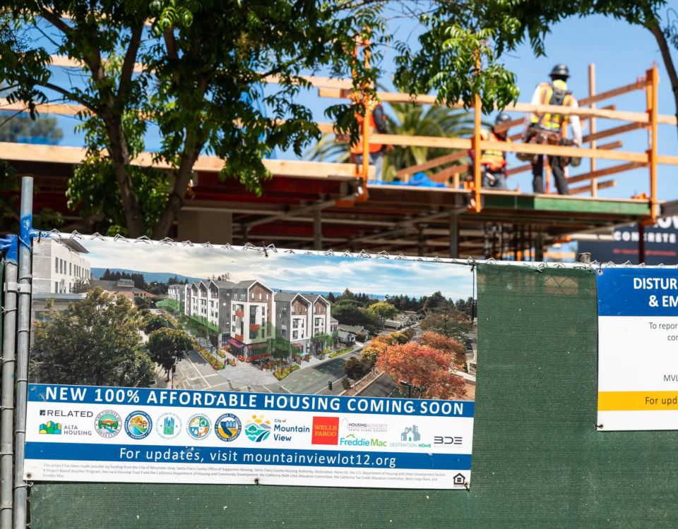 Green fence with sign that signifies affordable housing is being built. Workers in the background out of focus